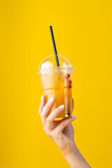 Summer Drink Fresh Lemonade with orange in the plastic cup on yellow background. female hand holding a glass. Mockup for your advertisement.