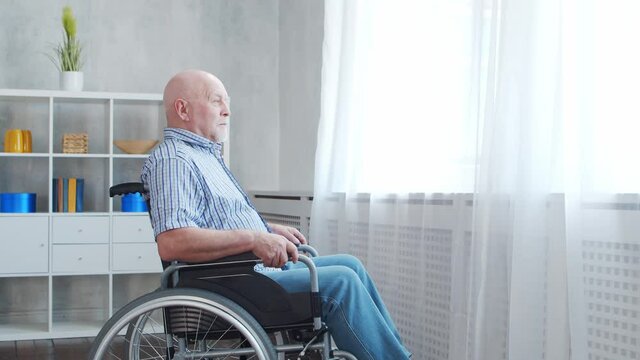 Disabled old man is sitting in a wheelchair at home alone. A handicapped person in a wheelchair is looking out the window. Paralysis and disability.