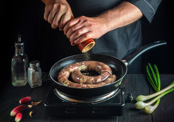 Chef or cook adds pepper to pan with raw meat sausage. Preparation for cooking sausages in kitchen of a restaurant or cafe on table with vegetables and spices.