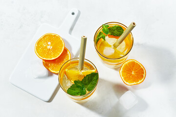 Top view of prepering summer cockteil. Glasses with gin and cold tonic with orange and mint, decorated with slices and with ice cubes on white background and bamboo straw.