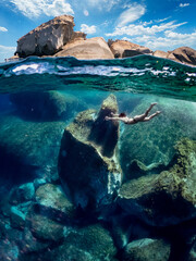 Young girl swimming underwater in deep water. Woman in half underwater effect while she swims surrounded by huge rocks.