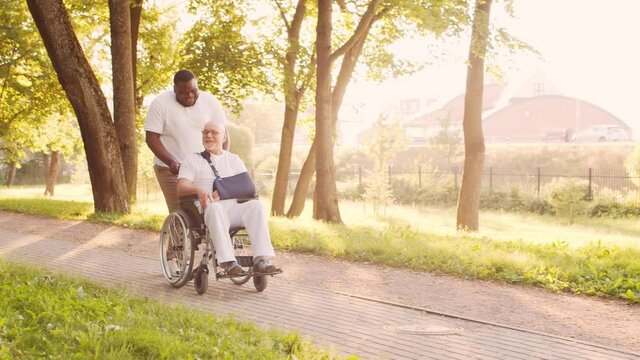 African-American caregiver and old disabled man in a wheelchair. Professional nurse and handicapped patient in the park. Assistance, rehabilitation and health care. Golden hour sunset.
