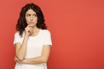 Doubtful young woman touching her chin, skeptic and pensive think with folded arms looking up to copy space over red studio background. Confident serious girl ponder on question or solution - obrazy, fototapety, plakaty