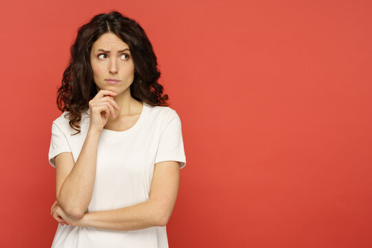 Doubtful young woman touching her chin, skeptic and pensive think with folded arms looking up to copy space over red studio background. Confident serious girl ponder on question or solution