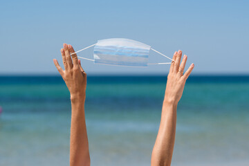 A blue medical mask is pulled by the hands of a girl on the beach while on vacation against the background of the sea.