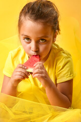 Caucasian little girl tasting watermelon and looking at camera. Yellow background. Fresh and healthy fruit concept.