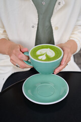 Woman with a cup of matcha latte sits on the summer terrace of a cafe. Delicious green matcha tea in a green cup close-up. Japanese hot milky beverage. Healthy Matcha Tea Made with Organic Ingredients