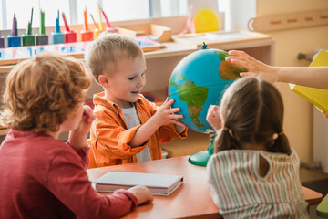 amazed boy looking at globe near blurred teacher and kids in montessori school