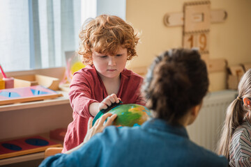 curly boy looking at globe near blurred teacher and girl in montessori school