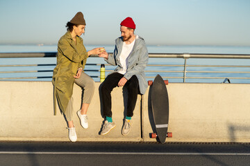 Happy couple drinking hot tea outdoors sitting on bridge broadwalk dressed in trendy street fashion...