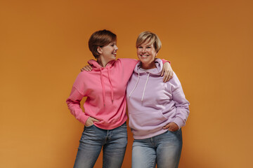 Joyful young brunette lady in pink stylish hoodie and jeans smiling and hugging old woman in lilac...