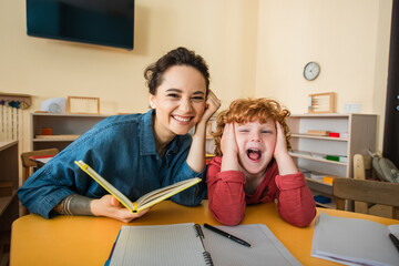 excited kid with open mouth touching head near cheerful teacher with book