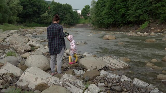 Little preschool children with mom on mountain river. Kids in raincoat throw stone to river. young woman take picture by smartphone and gimbal. Vacation, heading to camp, wild nature, family concept