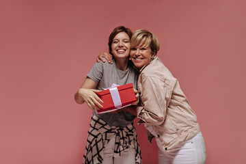 Happy two women with short cool hairstyle in white pants hugging, smiling and posing with red gift box on pink background..