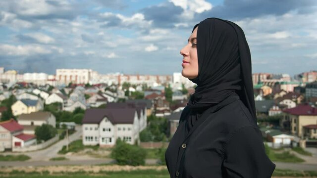 Pleasant Muslim woman in traditional black headscarf standing on mountain peak over Islamic city 