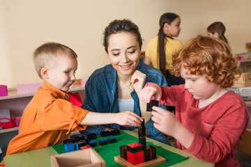 young teacher smiling near boys playing with multicolored cubes and interracial girls on blurred...