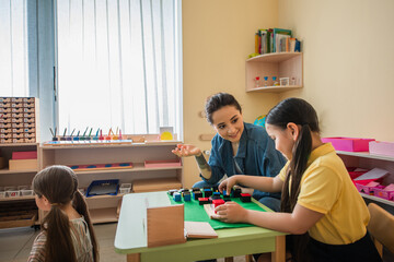 smiling teacher gesturing while playing multicolored cubes game with asian girl