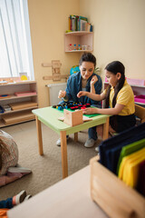 asian girl playing wooden cubes game with teacher near kids on floor in classroom