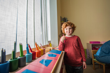 redhead boy looking at camera near color pencils in montessori school
