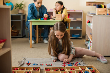 girl on floor playing with wooden letters near asian girl and teacher on blurred background