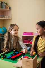 happy interracial girls looking at each other while playing with wooden blocks in montessori school