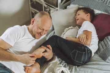 Fototapeta premium Father plays with his son 7-10 on bed. Dad tickles kids feet. Family, having fun