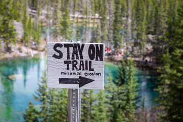 Wooden Sign with a right pointing arrow and Stay on Trail Minimize Erosion text on the Grassi Lakes Hike with blurred trees and turquoise lake in the background