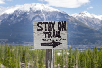 Wooden Sign with a right pointing arrow and Stay on Trail Minimize Erosion text on the Grassi Lakes Hike with blurred trees and mountains in the background