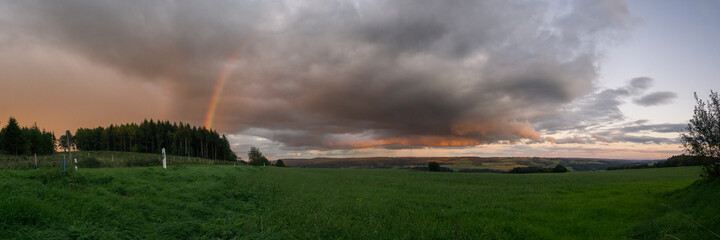 Naklejka na ściany i meble Panorama of a rainbow at sunset with red clouds over countryside with green fields, Eifel, Germany