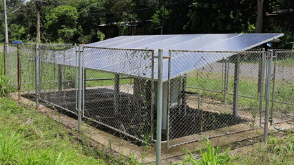 Solar panels and old metal fence. Clean energy panel for electricity generation with anti-theft steel grid fence in rural area on paved road background and green trees.