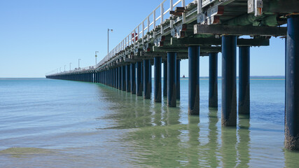 Perspective view of the Urangan Pier from the shoreline with gentle waves, blue sky and a distant sailing boat. 