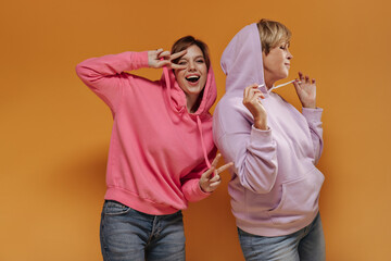 Cheerful young girl in pink sweatshirt showing peace signs, winking and posing with modern woman in lilac cool hoodies on orange backdrop..