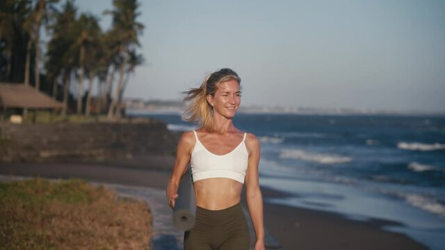 Attractive woman holding yoga mat walking along shore breathing fresh air, Bali