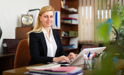 Middle age european businesswoman in business suit sitting in office at workplace and working at laptop