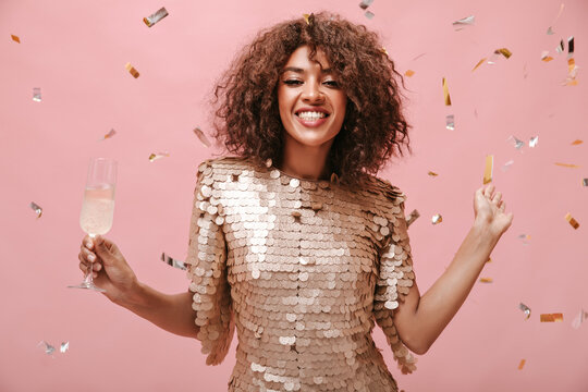 Positive brunette woman in modern beige dress smiling and posing with glass with wine and with confetti on pink backdrop..