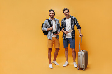 Happy young good-humored men in checkered shirts and colorful shorts smile and pose on orange background. Travelers hold tickets, retro camera and suitcase.