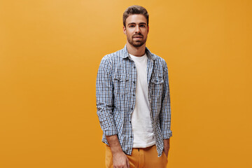 Serious young bearded man in orange pants, stylish checkered shirt and white t-shirt looks into...