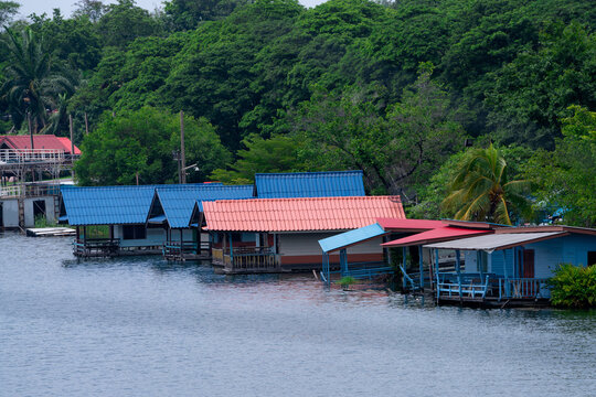 A house built on the waterfront in a rural area in Thailand. The house has a blue roof and a red roof, behind it is a big tree.