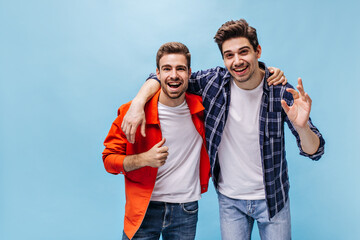 Charming man in orange jacket shows thumb up. Brunet guy in checkered shirt and his friend smile on blue background.