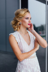 Beautiful blond bride in white wedding dress standing near mirror window outdoors.