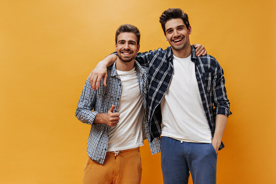 Charming men in checkered shirts and white t-shirt smile on orange background. Cool bearded guy shows peace sign.