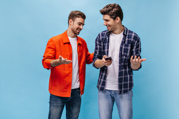Cheerful men talk on blue background and smile. Guy in checkered shirt holds phone. Friends pose on...