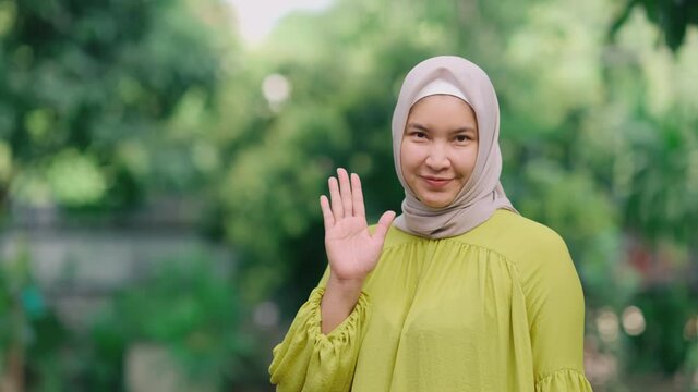 A beautiful middle-aged Asian Muslim woman. Standing smiling and raising a salam hand to greet the camera with bright cuteness at the garden.