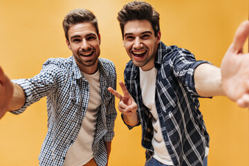 Happy men in checkered shirts, white t-shirts laugh on orange background and take selfie. Brunet...