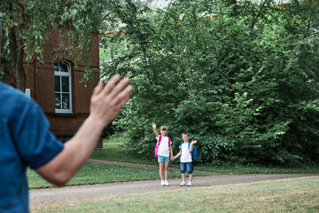 Obraz premium Dad takes the kids to school or kindergarten, stands and waves at them. Happy kids return to school after vacation. Morning rituals, school days