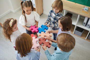 Team of primary school children or kindergartners standing in circle join pieces of colorful jigsaw puzzle, high angle close up. Education, learning, engaging activities in classroom, teamwork concept