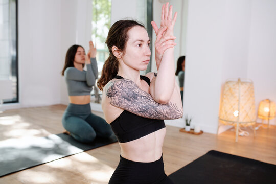 Conentrated women in sportswear practicing yoga in a group, meditating with arms interlaced in a big studio. Focus on a girl with big shoulder tattoo
