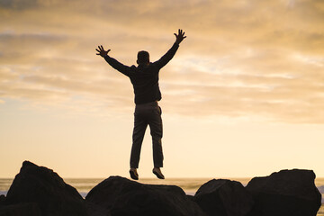 Fototapeta na wymiar happy jumping MAN in YES POSE on beach rocks AT THE ocean WITH arms wide open enjoying life 
