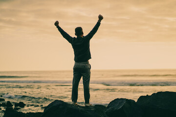 happy successful MAN  in YES POSE on beach rocks AT THE ocean WITH arms wide open enjoying life
