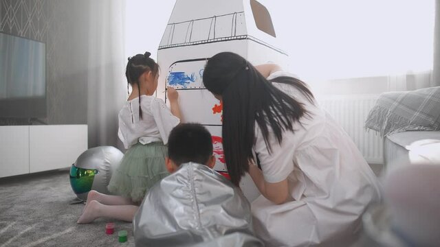 Handheld, an asian female with kids play in the living room at home, a boy in an astronaut costume sitting on the floor with her mother and sister, children together with their mother paint on a